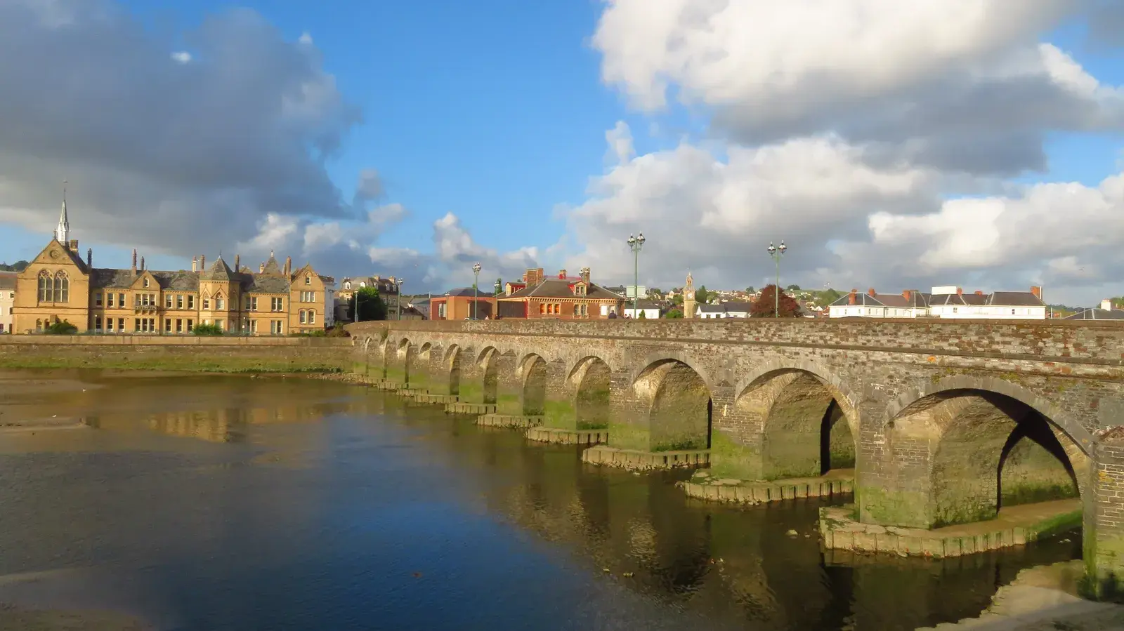 The stone arches of Barnstaple's Long Bridge reflected in the River Taw, with Victorian buildings on the far bank and white clouds in a blue sky.