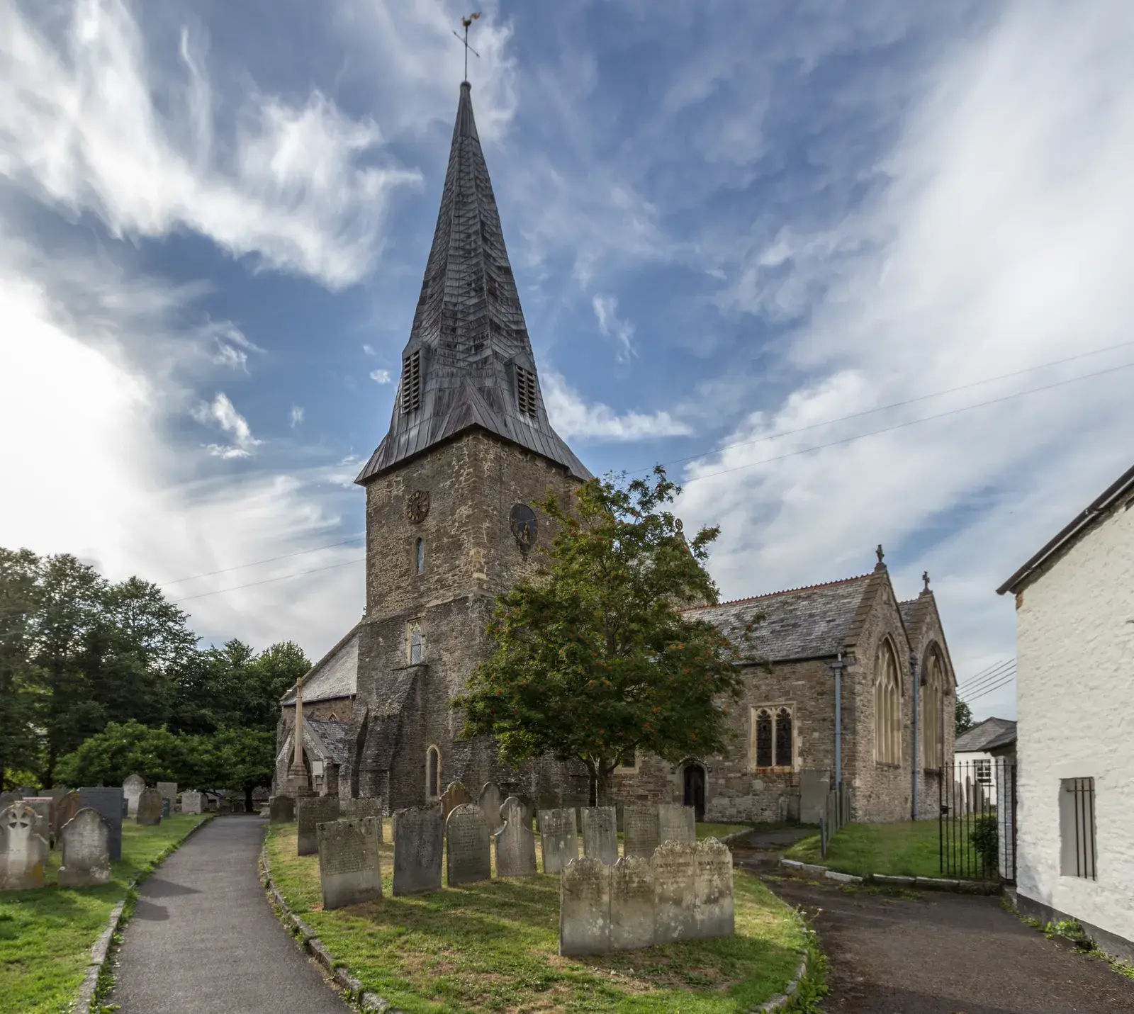 St Brannock's Church in Braunton: a stone tower with a tall lead-covered spire, the churchyard with old headstones and a path leading to it.