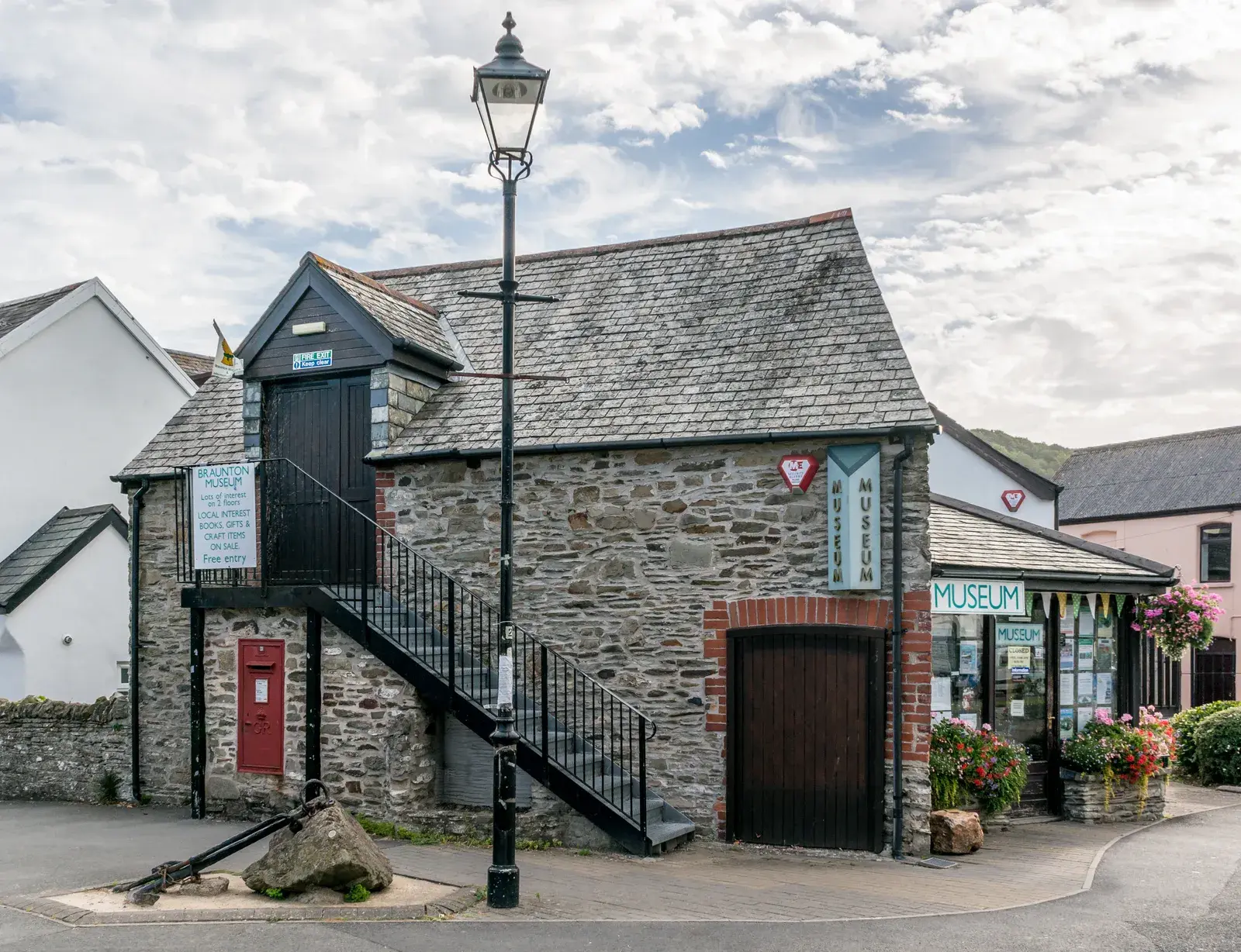 Braunton & District Museum in the village centre: a stone building with an outside staircase, a red post box, an old anchor and a street lamp in front, and flower baskets by the entrance.