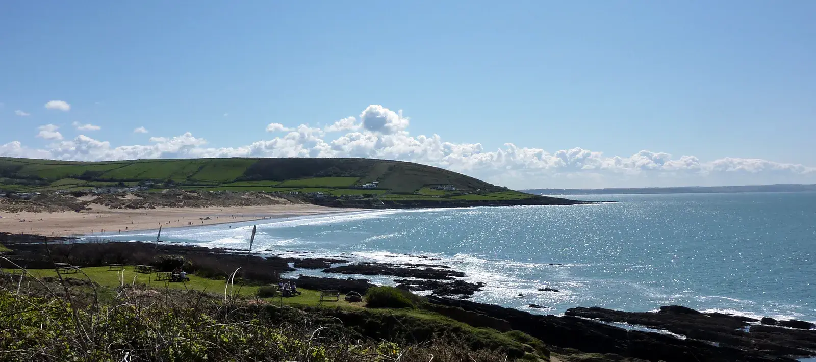 Croyde Bay on a sunny day, seen from the rocky headland: dark rocks and grass in the foreground, sparkling sea, the sandy beach and a green headland across the bay.
