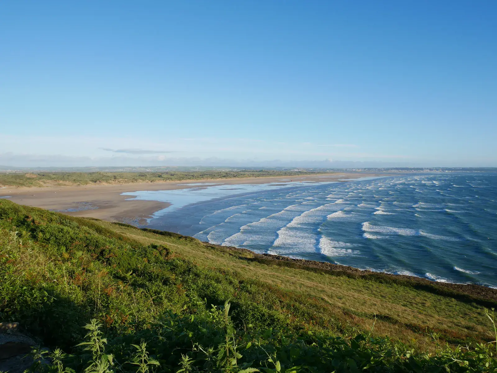 Saunton Sands seen from the grassy slope above its northern end on a clear day: rows of white surf rolling onto a long, curving beach with dunes behind.