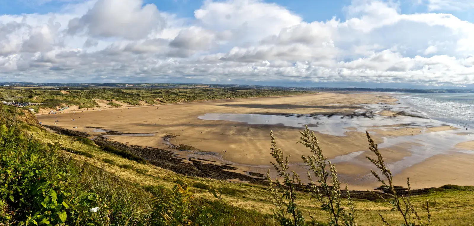 Wide panorama of Saunton Sands at low tide from the hillside: a broad sandy beach with shallow pools, grass-covered dunes stretching into the distance and white clouds overhead.