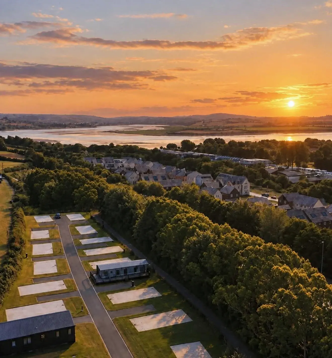 Drone photo of the park: the pitches and lane of Chivenor Park in the foreground, then trees, the houses of Chivenor and the Taw estuary, with the sky and colour edited to an evening glow.