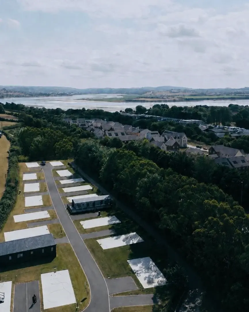 Drone photo in daylight looking along the lane and the pitches of Chivenor Park, with trees beside them and the Taw estuary beyond.